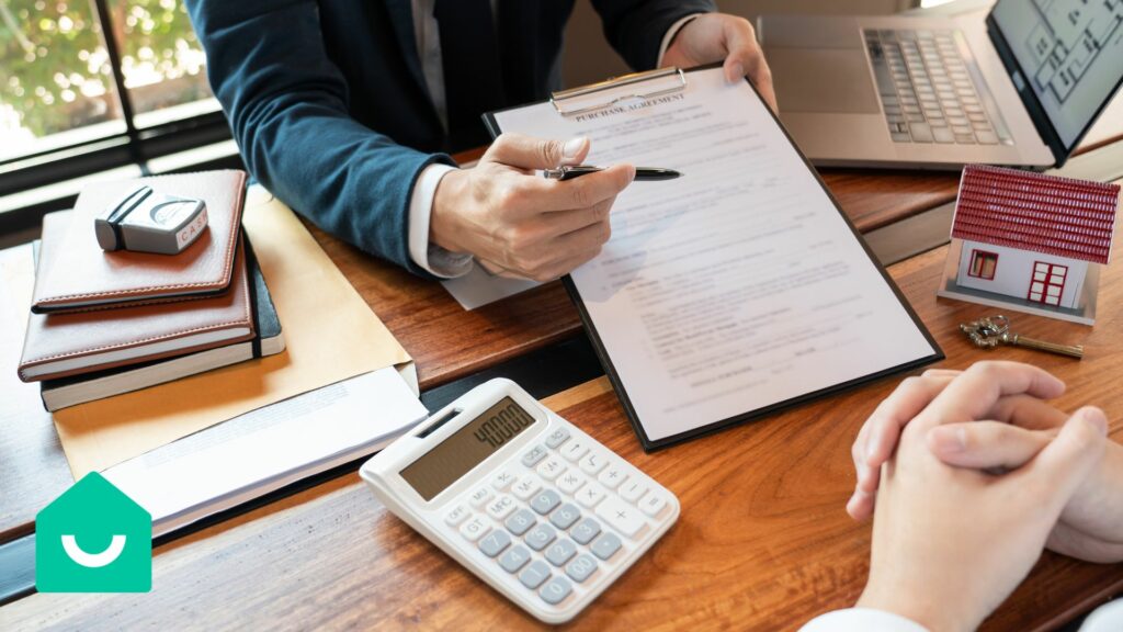 Un agent immobilier expliquant un contrat d'achat à un client, avec des dossiers, une calculatrice et une maquette de maison sur la table.