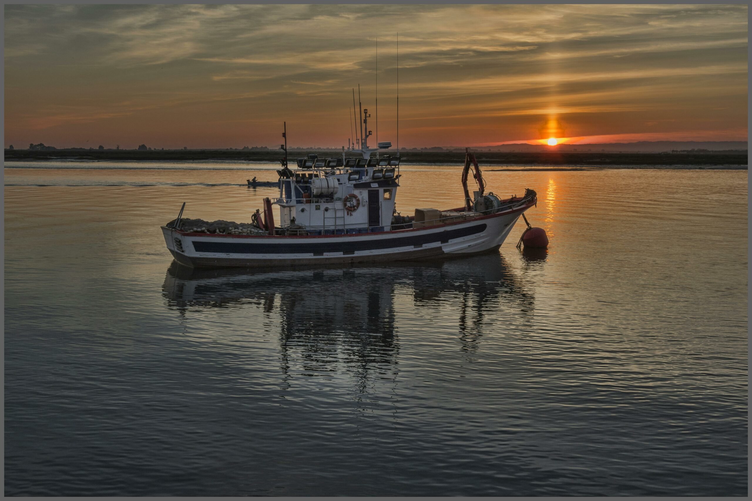 Un bateau de pêche ancré sur l'eau calme au crépuscule, avec le soleil se couchant à l'horizon et reflétant ses couleurs dorées sur la surface de l'eau.