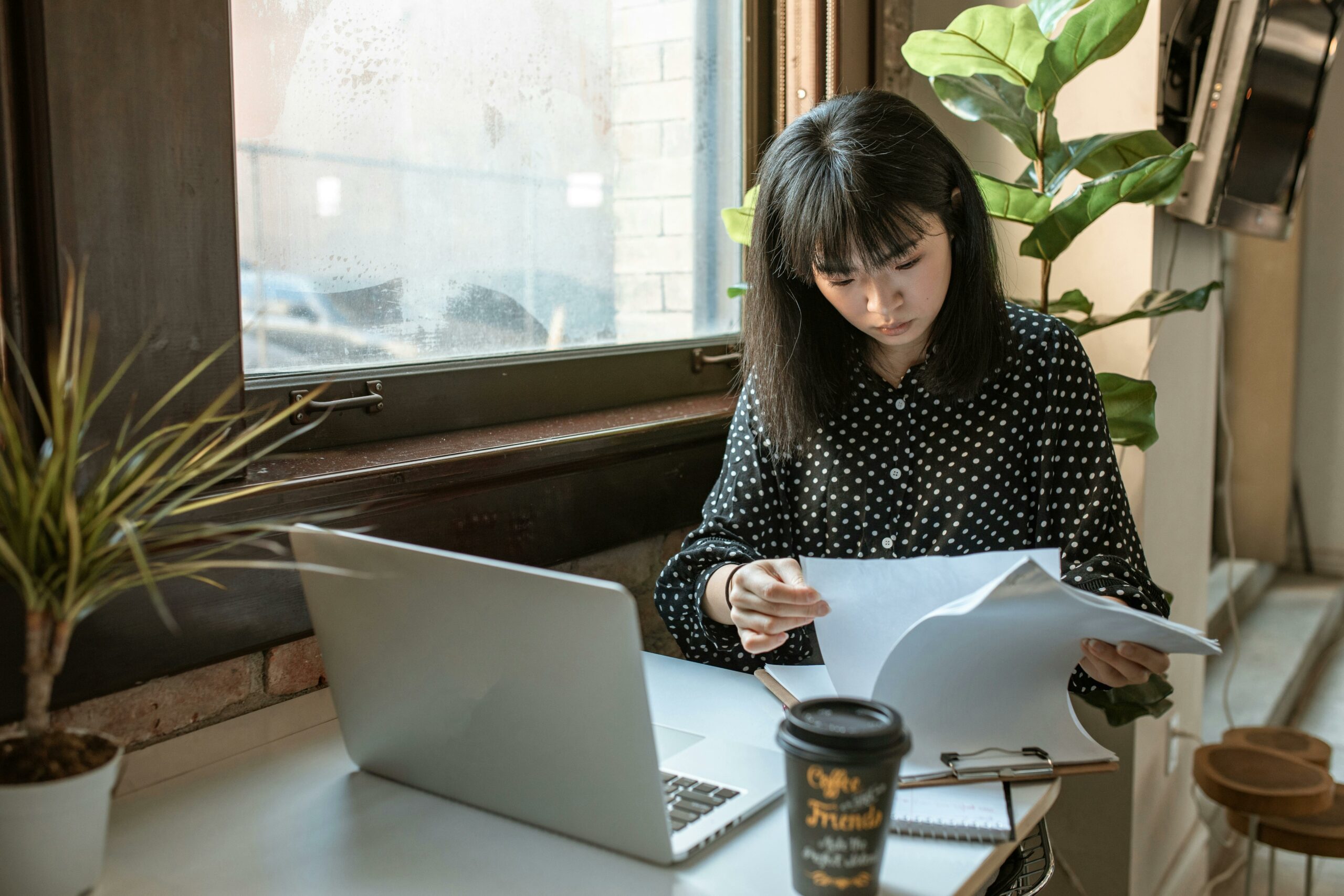 Une femme, assise à un bureau, examine des documents tout en travaillant sur un ordinateur portable, avec une plante en pot et un gobelet de café à côté, dans un environnement lumineux près d'une fenêtre.