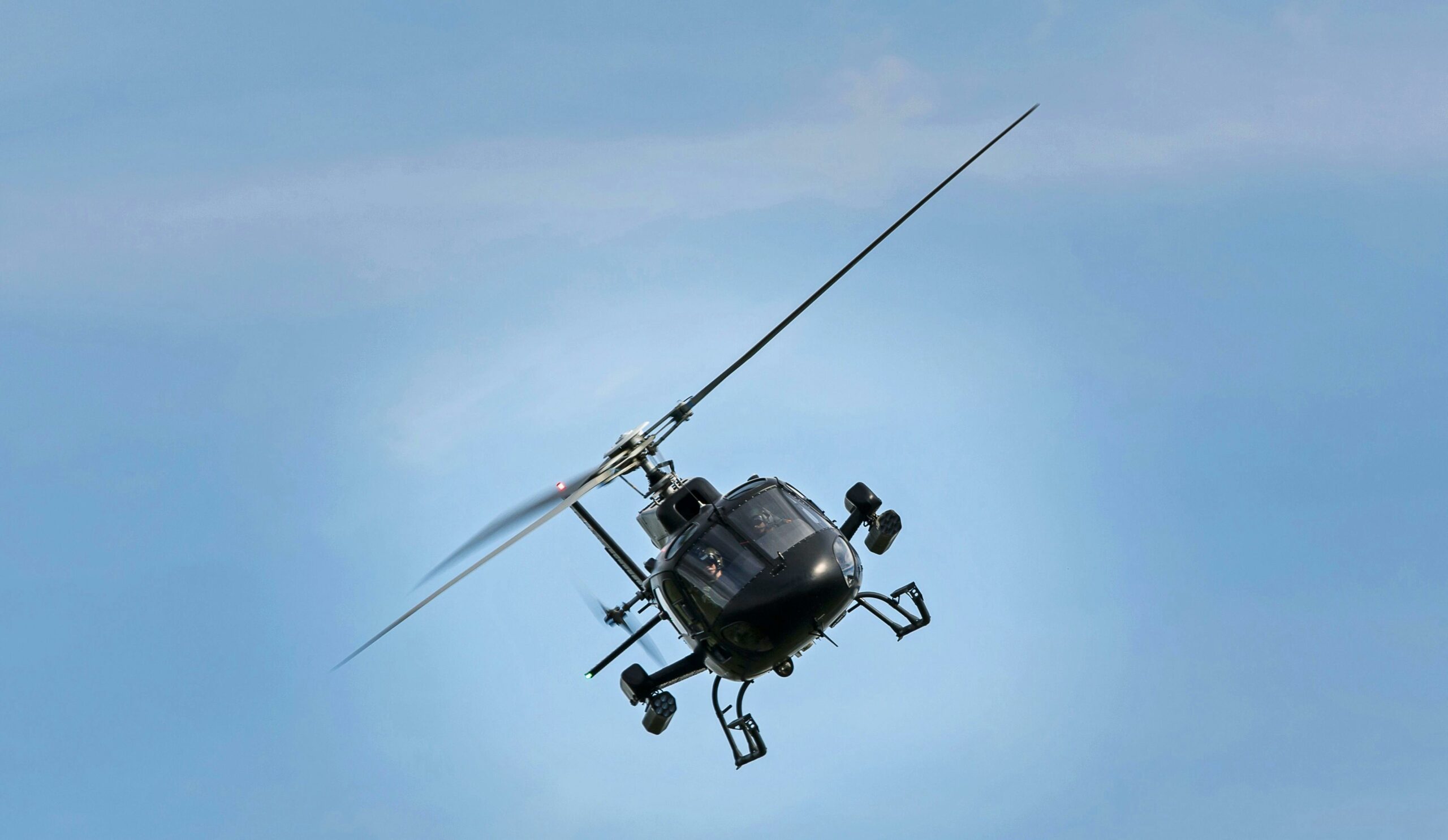 Un hélicoptère noir en vol incliné contre un ciel bleu clair, vu de face avec ses pales en mouvement rapide.