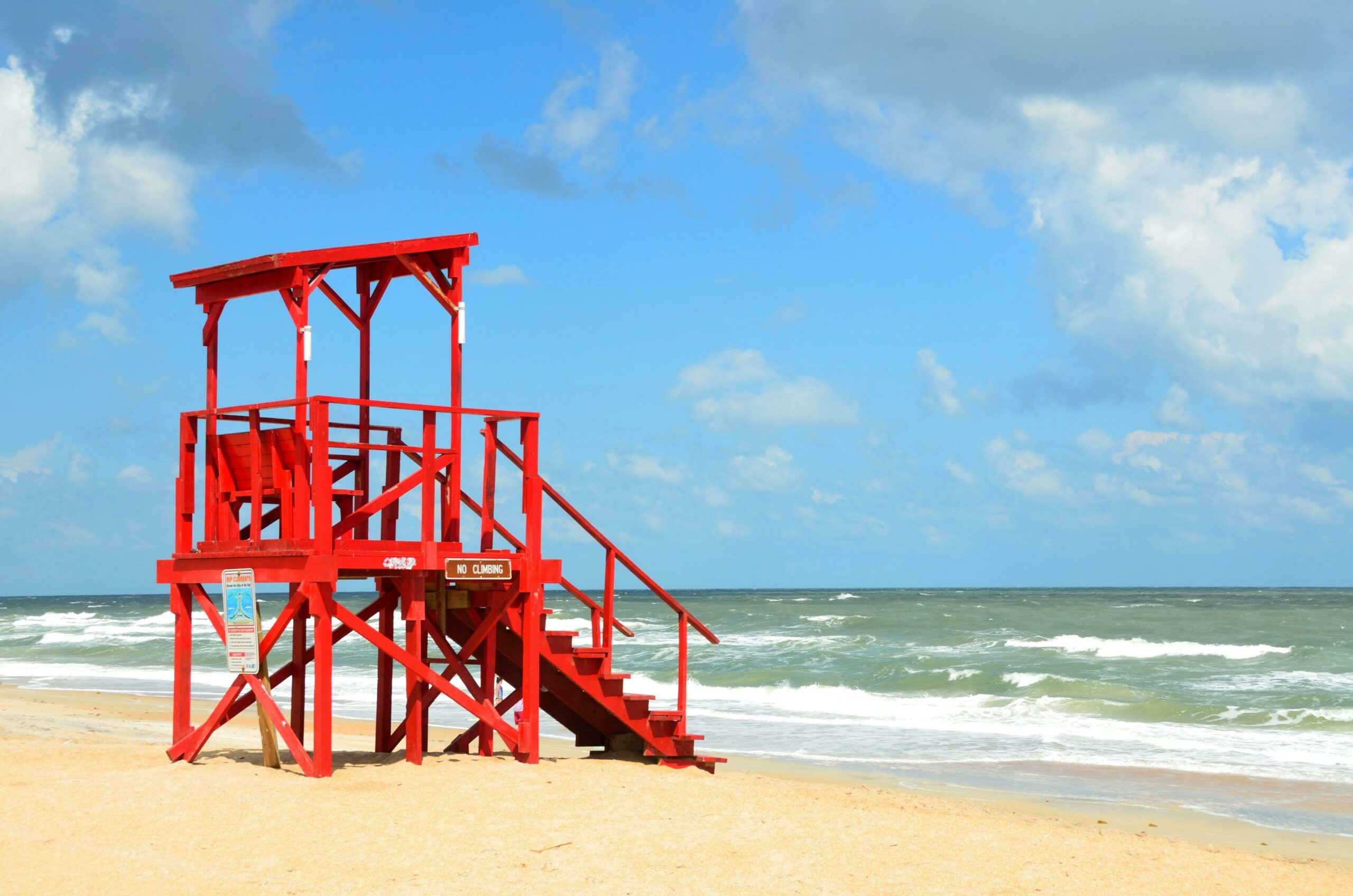 Un poste de surveillance de plage rouge vif sur le sable, avec l'océan et un ciel partiellement nuageux en arrière-plan.