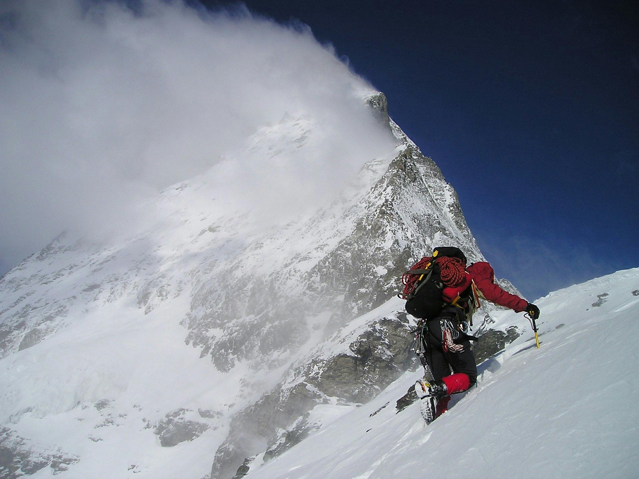 Un alpiniste en tenue rouge et noire escaladant une montagne enneigée avec un piolet, sous un ciel bleu avec des nuages se formant autour du sommet.