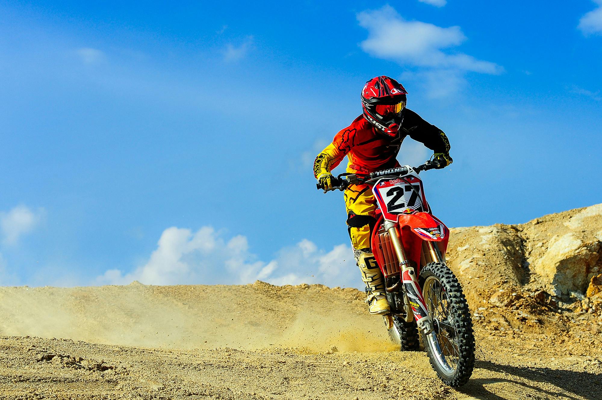Un motocycliste en tenue rouge et jaune, portant le numéro 27, pilotant une moto de cross rouge sur une piste sablonneuse sous un ciel bleu dégagé.