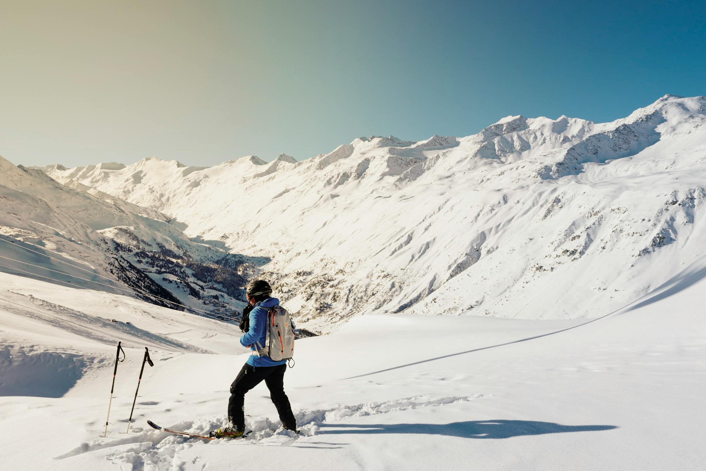 Un skieur en tenue bleue et noire, portant un sac à dos, se tenant sur une piste enneigée avec des montagnes majestueuses en arrière-plan sous un ciel bleu dégagé.