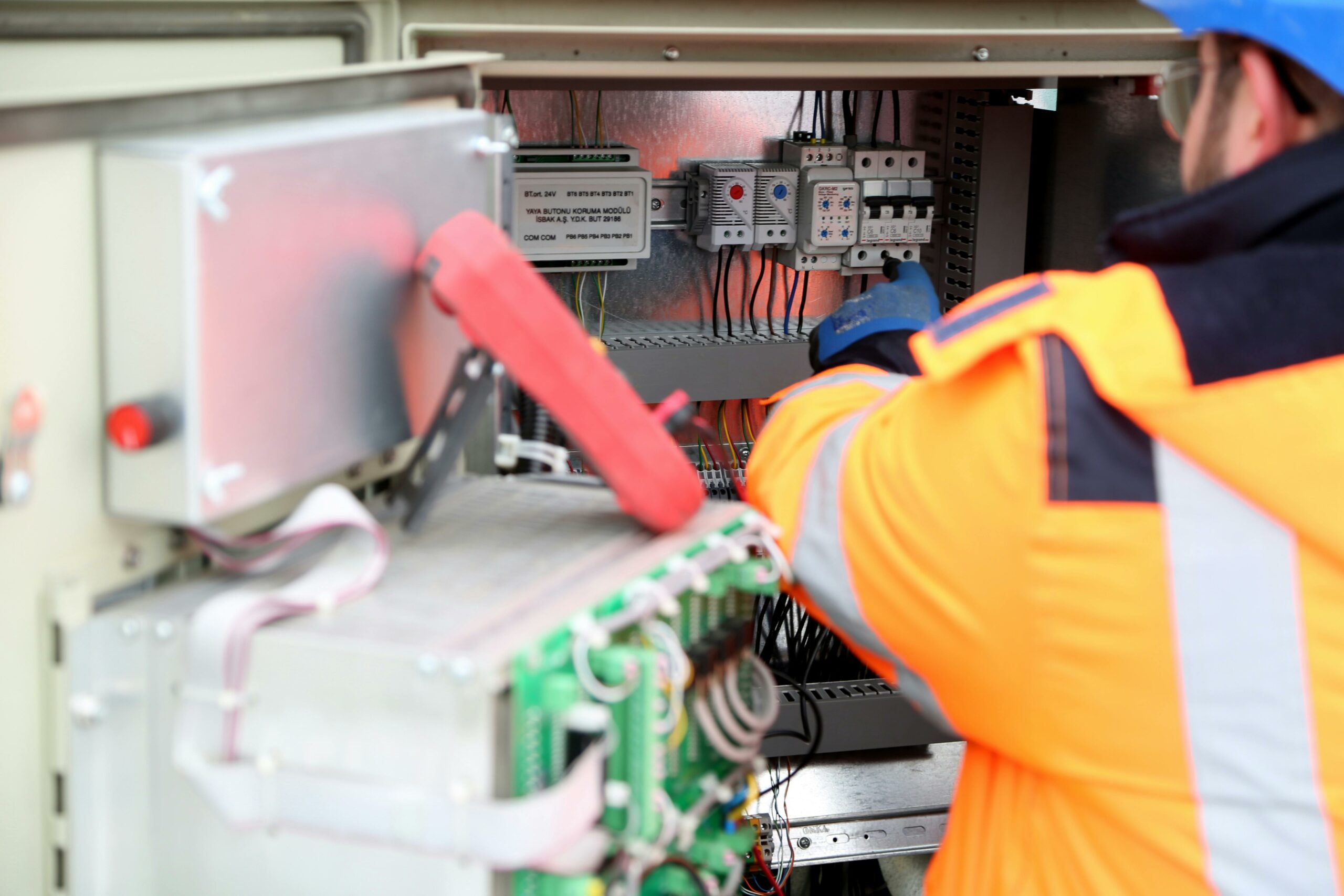 Un technicien en veste orange haute visibilité et casque bleu travaille sur un panneau électrique ouvert, ajustant des composants à l'intérieur.