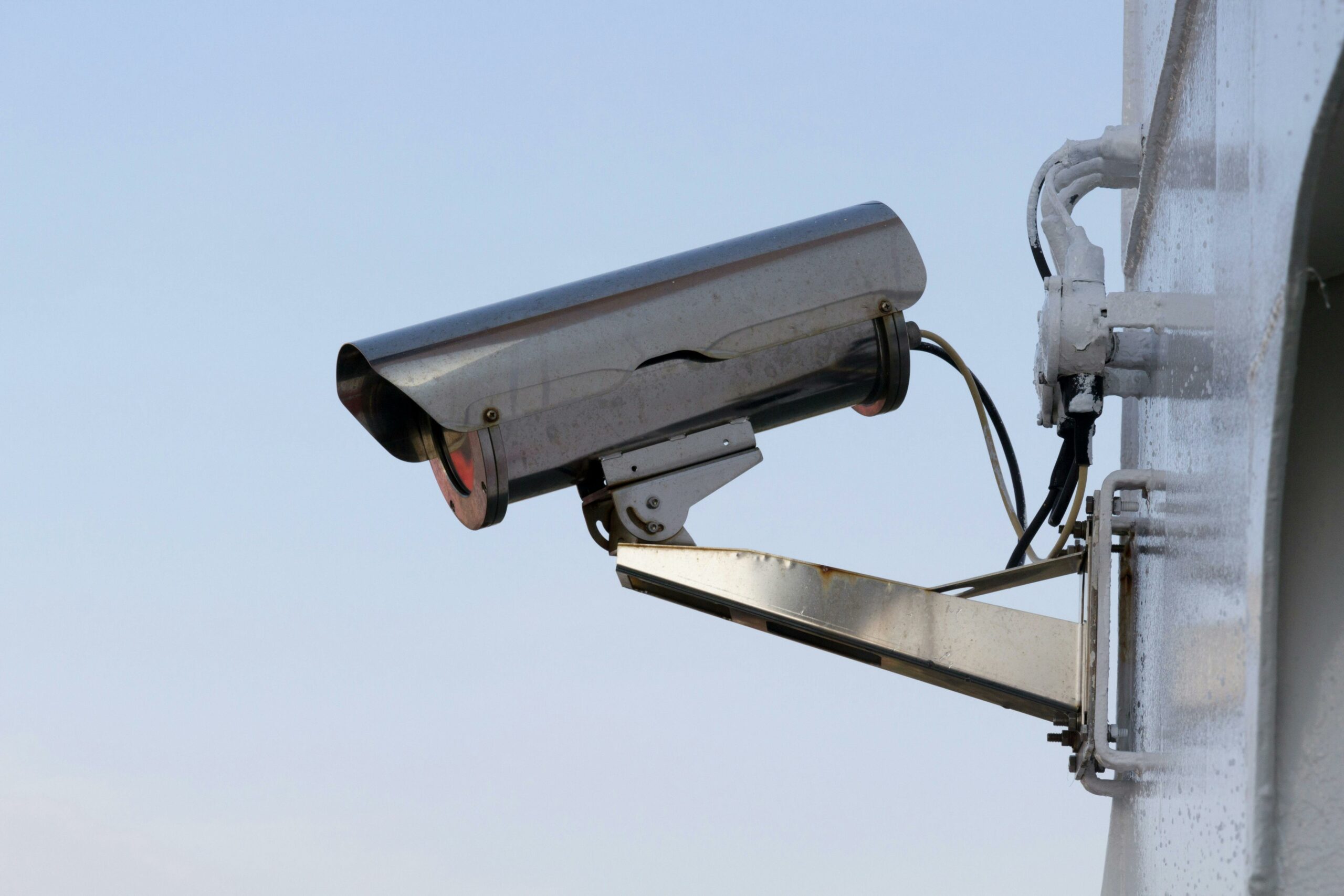 Une caméra de surveillance en métal fixée sur un mur, pointée vers la droite, avec un ciel bleu clair en arrière-plan.