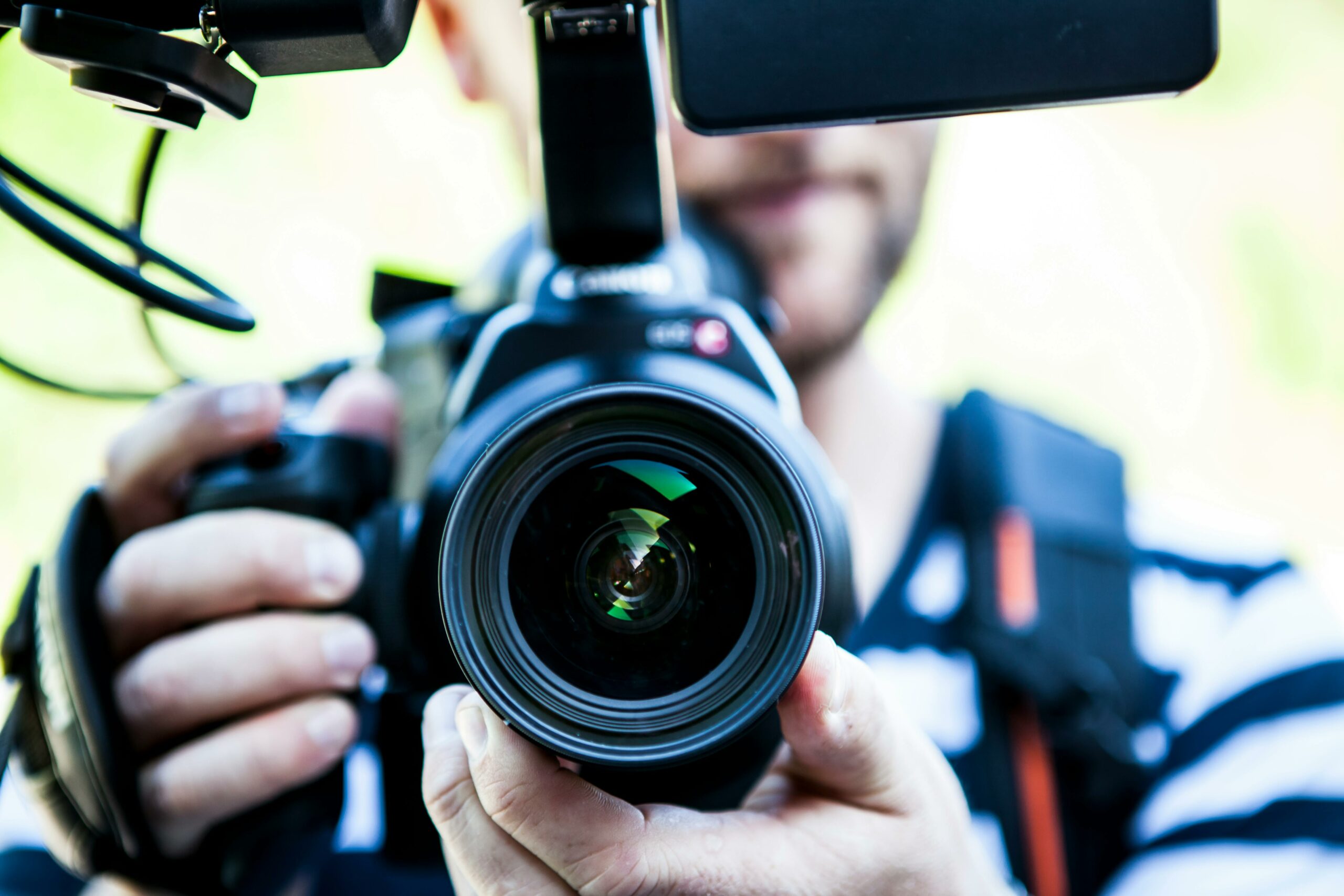 Gros plan sur une caméra tenue par un vidéaste, avec l'objectif en focus. On aperçoit en arrière-plan flou une personne en tenue rayée.