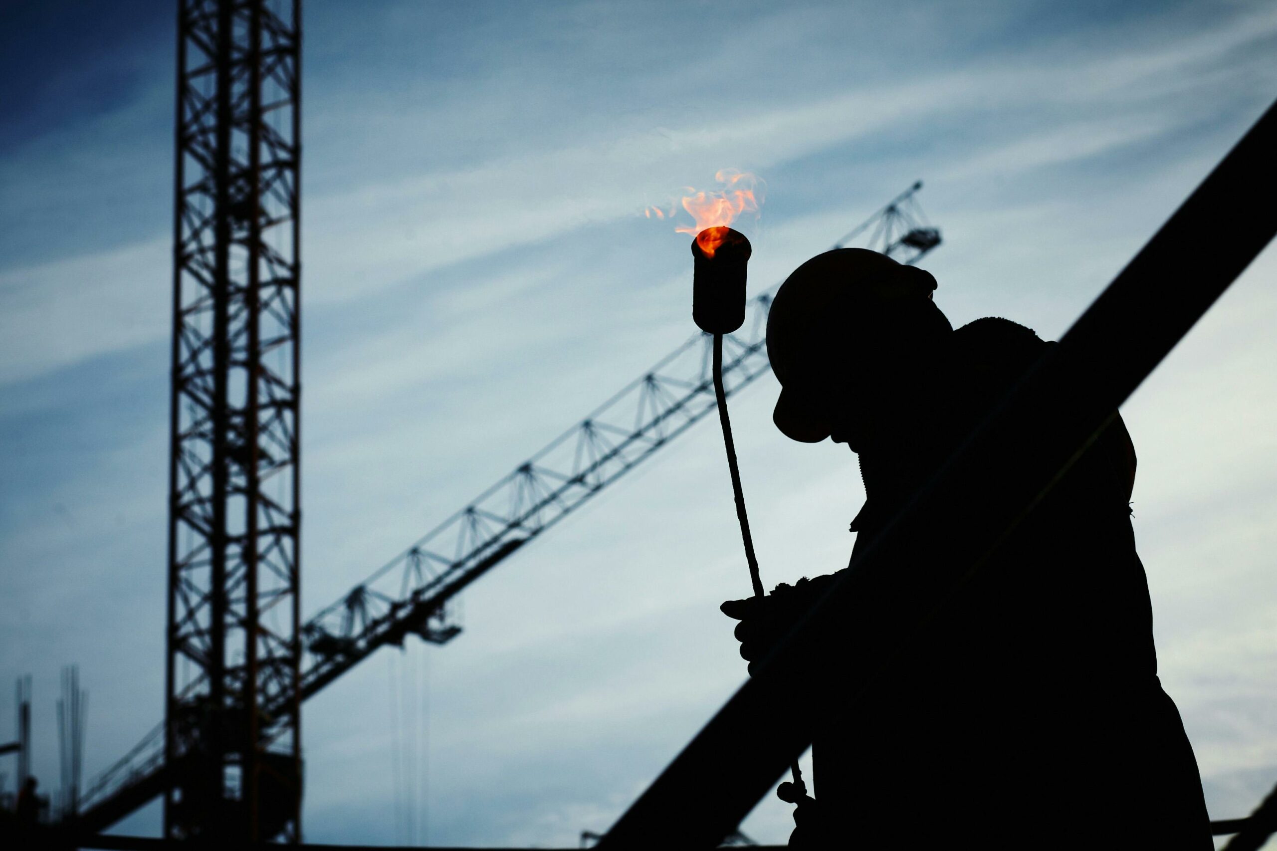 Silhouette d'un ouvrier tenant une torche allumée avec des flammes, se détachant sur un ciel bleu avec des grues en arrière-plan.